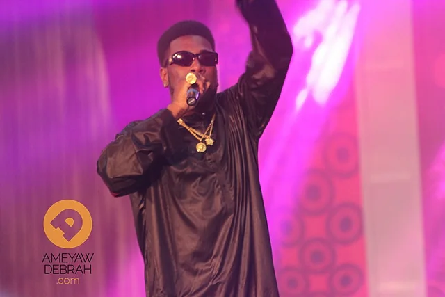 Burna Boy performs into a microphone under pink stage lighting.