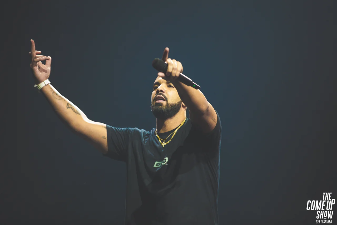 Drake onstage in a black T-shirt, holding a microphone with both arms raised